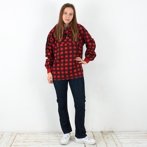 May include: A person wearing a red and black plaid shirt with a zipper neckline and dark blue jeans. The shirt has long sleeves with red cuffs. The person is wearing white sneakers.