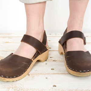 Może przedstawiać: Para brązowych drewniaków ze skórzaną cholewką i drewnianą podeszwą oraz paskiem na górze stopy i wokół kostki. Drewniaki mają zaokrąglony nosek i mały obcas. Buty są pokazane na białej drewnianej powierzchni.