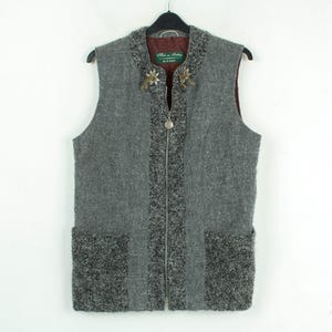 Puede incluir: Un chaleco de lana gris con cremallera y dos bolsillos delanteros. El chaleco tiene un diseño texturizado con un ribete gris más oscuro y alfileres florales decorativos cerca del cuello. El chaleco está colgado de una percha negra.