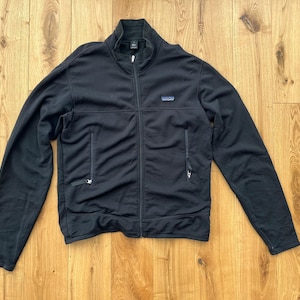 May include: Black zip-up jacket with a stand-up collar and two side pockets. The jacket has a small blue and white logo patch on the chest. The jacket is made of a soft, textured material.
