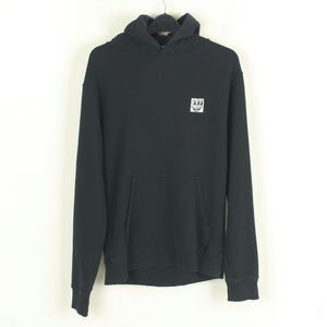 May include: Black hooded sweatshirt with a kangaroo pocket and a small white emblem on the chest. The sweatshirt is hanging on a black hanger against a white background. The hoodie has long sleeves and a drawstring hood.