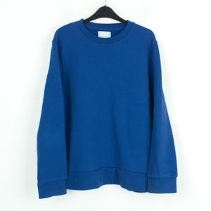 May include: A solid blue sweatshirt with a crew neck and long sleeves. The sweatshirt is hanging on a black hanger against a white background. The cuffs and hem are a darker shade of blue. A small white tag is visible on the neckline.