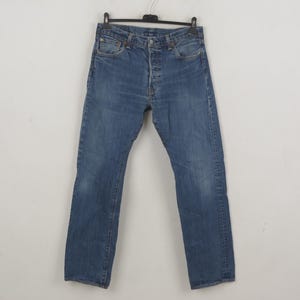 Puede incluir: Vaqueros de mezclilla azules colgados de una percha negra. Los vaqueros tienen una bragueta con botones y un diseño clásico de cinco bolsillos. La mezclilla tiene un aspecto descolorido, con zonas más claras en los muslos y las rodillas.