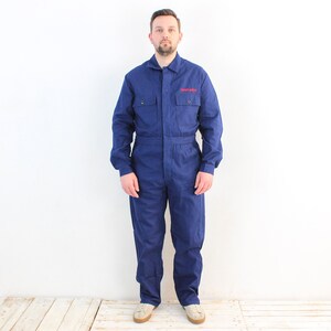 May include: A navy blue work jumpsuit with a collared neckline and long sleeves. The jumpsuit has two chest pockets and the word "Snap-on" in red lettering on the left chest. The garment is worn by a person.
