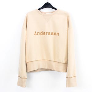 May include: A beige sweatshirt with the embroidered word "Andersson" in a brown thread. The long-sleeved top has a crew neck and ribbed cuffs and hem. The sweatshirt is hanging on a black hanger against a white background.