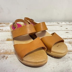 Può includere: Un paio di sandali in pelle marrone chiaro con suole in legno. I sandali hanno due cinturini sulla parte superiore del piede e un cinturino alla caviglia regolabile. Le suole in legno sono spesse circa 2,5 cm. Sfondo in legno verniciato di bianco.