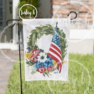 May include: A garden flag featuring a watercolor design of an American flag, a floral wreath, and the text "haley b designs". The flag is displayed on a black metal stand, set against a backdrop of green grass.