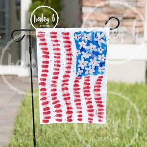 May include: A white garden flag with a painted American flag design. The flag features red stripes and a blue square with white flower accents. The flag is displayed on a black metal stand in a grassy yard.