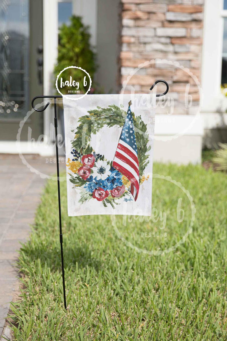 May include: A garden flag featuring a watercolor design of an American flag, a floral wreath, and the text "haley b designs". The flag is displayed on a black metal stand, set against a backdrop of green grass.