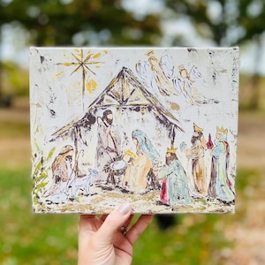 Puede incluir: Un lienzo pintado a mano que representa la escena de la Natividad con un establo de madera r&uacute;stico, una estrella, &aacute;ngeles y tres reyes magos. La escena est&aacute; pintada en un estilo caprichoso con colores apagados.