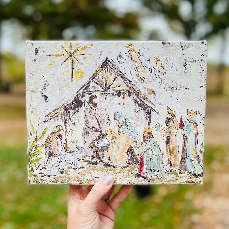Puede incluir: Un lienzo pintado a mano que representa la escena de la Natividad con un establo de madera r&uacute;stico, una estrella, &aacute;ngeles y tres reyes magos. La escena est&aacute; pintada en un estilo caprichoso con colores apagados.