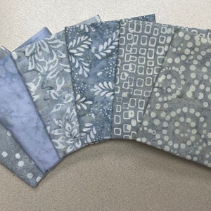 May include: A collection of seven fabric squares in shades of blue and gray, arranged in a fan shape. Each square features a unique pattern, including dots, squares, and floral designs. The fabric appears to be cotton or a similar material, suitable for quilting or crafting projects.