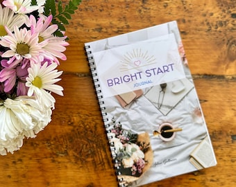 May include: A white and pink floral bouquet sits on a wooden table next to a spiral bound journal with the title "BRIGHT START JOURNAL" on the cover.