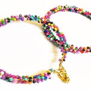 Pode incluir: Duas pulseiras de contas coloridas com fechos dourados. As pulseiras são feitas de contas pequenas multicolores, incluindo roxo, rosa, laranja, amarelo, verde, azul, preto e branco.