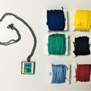 Puede incluir: Collar con colgante cuadrado con diseño de punto de cruz con detalles azules, verdes y rojos. La cadena es oscura. Cerca se disponen carretes de hilo de bordar azul, amarillo, verde, negro, azul claro y rojo.
