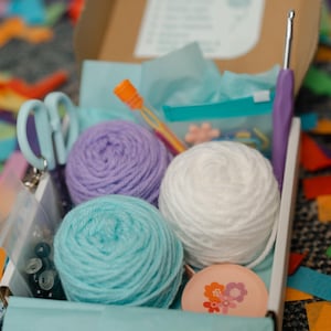 May include: A craft box filled with yarn, crochet hook, scissors, and other supplies. The yarn is in shades of purple, white, and teal. The box is open, revealing the contents, and is surrounded by colorful confetti.