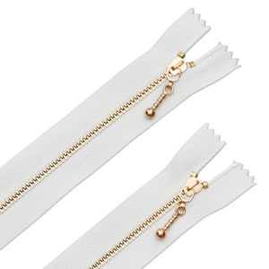 Könnte beinhalten: Zwei weiße Reißverschlüsse mit goldenen Metallzähnen und -ziehern. Die Zipper haben eine kleine, runde, goldene Perle am Ende.