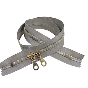 May include: A coiled gray zipper featuring two gold-toned metal sliders with circular pull tabs. The zipper tape is a solid gray fabric, and the teeth are metallic silver. The two sliders are positioned side-by-side on the zipper track.
