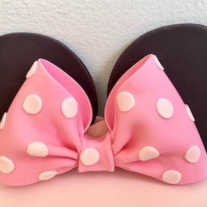 May include: Pink bow with white polka dots, attached to two black ears. The bow is made of fondant and is a popular decoration for cakes.