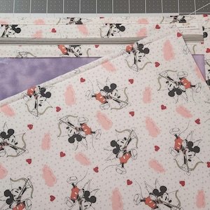 May include: A white fabric pouch with a zipper closure and a clear plastic window. The fabric features a repeating pattern of pink hearts and Mickey Mouse characters dressed as cupids with pink wings and holding arrows.