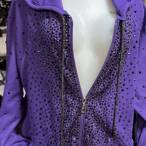 Peut inclure: Un sweat-shirt à capuche violet avec fermeture éclair orné de nombreux ornements de couleur foncée. Le sweat-shirt est doté d'une fermeture éclair intégrale, d'une capuche et de poches. Les ornements sont concentrés autour de la fermeture éclair.