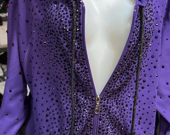 May include: A purple hooded zip-up sweatshirt adorned with numerous dark-colored embellishments. The sweatshirt features a full zipper, a hood, and pockets. The embellishments are concentrated around the zipper and spread across the front.
