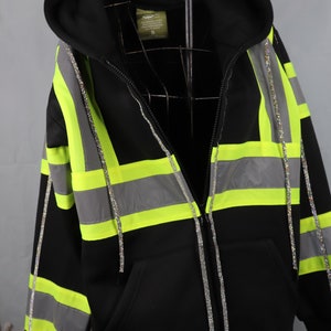 Può includere: Felpa con cappuccio nera con strisce riflettenti argentate e verde neon. La felpa ha una chiusura a zip ed è decorata con una linea di strass su ogni lato.