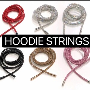 May include: Six pairs of sparkly hoodie strings in various colors, including red, silver, black, gold, pink, and clear. The strings are made of rhinestones and are tied in a knot at the ends. The text "HOODIE STRINGS" is displayed in the center of the image.