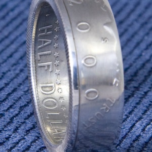 May include: A silver half dollar coin ring with the words "HALF DOLLAR" and "IN GOD WE TRUST" engraved on the side. The coin is dated "2000".