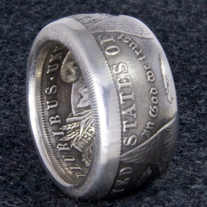 May include: A silver ring made from a US silver dollar coin. The ring features the words "United States of America" and "In God We Trust" on the side. The ring is on a dark gray fabric background.
