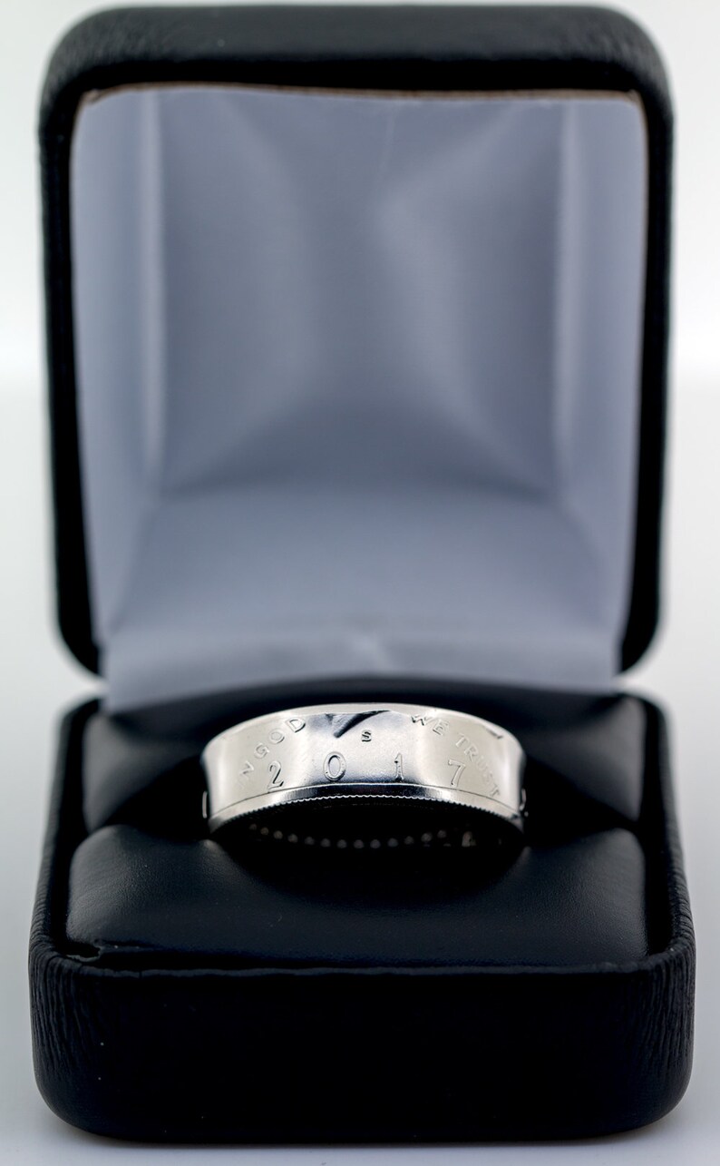 May include: A silver coin ring with the inscription "In God We Trust" and the year "2017" on the band. The ring is displayed in a black velvet ring box.