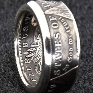 Puede incluir: Anillo de plata elaborado a partir de una moneda, con inscripciones como "In God We Trust" y "United States of America". El anillo tiene una superficie texturizada y un acabado pulido, mostrando el dise&ntilde;o original de la moneda. Una pieza de joyer&iacute;a &uacute;nica.