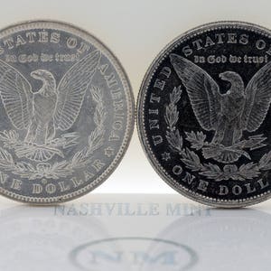 Puede incluir: Dos monedas de plata Morgan. Las monedas presentan un &aacute;guila con las alas extendidas y el texto "STATES OF AMERICA" y "ONE DOLLAR". Tambi&eacute;n son visibles las palabras "In God We Trust". Las monedas se muestran sobre una superficie blanca.
