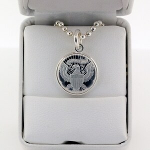 May include: A silver necklace with a round pendant featuring the Presidential Seal of the United States. The pendant is in a white box with a white interior.