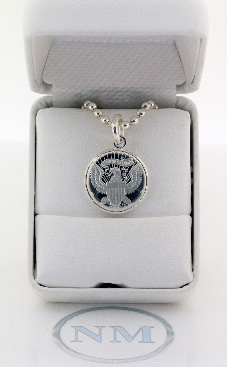 May include: A silver necklace with a round pendant featuring the Presidential Seal of the United States. The pendant is in a white box with a white interior.