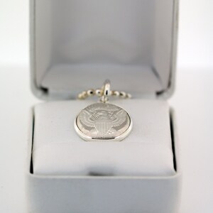 May include: A silver pendant necklace with a round, engraved charm featuring the Presidential Seal of the United States. The necklace is displayed in a white box.