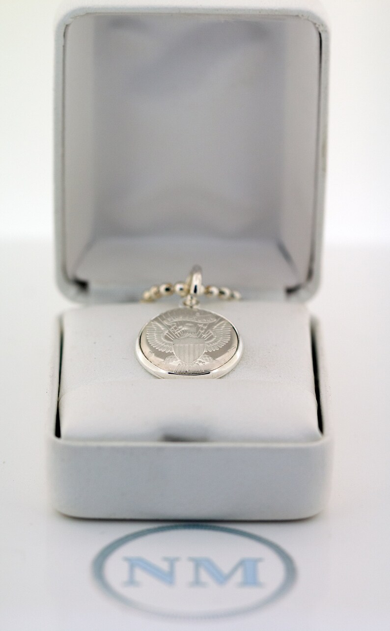 May include: A silver pendant necklace with a round, engraved charm featuring the Presidential Seal of the United States. The necklace is displayed in a white box.