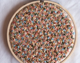 May include: A close-up of a circular embroidery hoop with a colorful, textured design. The design is made up of small, tightly packed loops of thread in shades of pink, blue, green, and brown.
