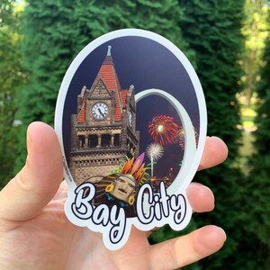 May include: A round sticker with a colorful illustration of a building with a clock tower, fireworks, and a Native American headdress. The text "Bay City" is written in a stylized font below the illustration.