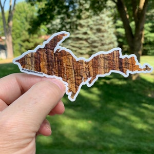May include: A sticker of the Upper Peninsula of Michigan, shaped like the peninsula, with a brown and tan wood grain pattern. The sticker has a white border and the text "©CraigSterken.com" in the lower left corner.