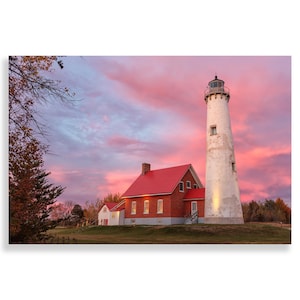 May include: A white lighthouse with a red roof stands tall against a pink and purple sunset sky. A smaller red brick building with a red roof is in the foreground.