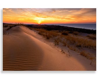 May include: A large sand dune with a rippled surface, illuminated by the warm glow of a sunset over a body of water. The sky is a mix of orange, pink, and purple hues.