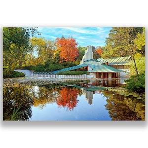 May include: A modern, white house with a green roof sits on a pond surrounded by trees. The trees are in various shades of green, yellow, and orange, indicating autumn. The house is reflected in the still water of the pond.