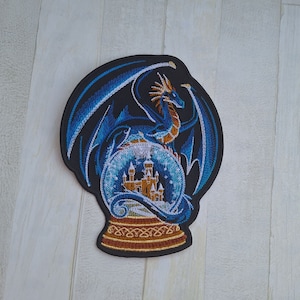 Può includere: Una toppa ricamata con un drago blu avvolto attorno a una sfera di neve che contiene un castello dorato. La sfera di neve è su uno sfondo nero.