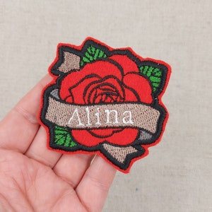 Può includere: Rosa rossa ricamata con foglie verdi e uno stendardo che dice "Alina" in filo bianco. La rosa è in stile tradizionale di tatuaggio.