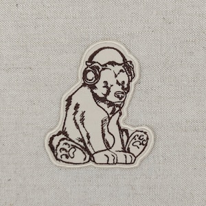 Puede incluir: Parche bordado con un oso sentado con auriculares. El oso está representado en un contorno marrón con un relleno color crema. El parche está sobre un fondo de tela beige claro.