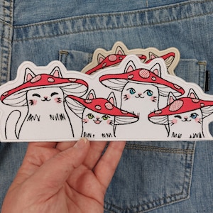 May include: Four white embroidered iron-on patches of cats wearing red mushroom hats. The patches are on a blue denim background.