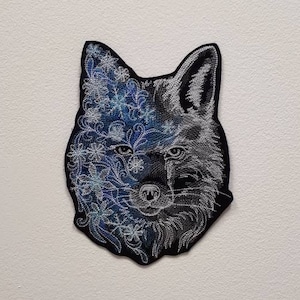 Pode incluir: Um remendo bordado em preto e branco de um rosto de lobo com um design floral de um lado. O rosto do lobo é detalhado com um nariz e olhos pretos.