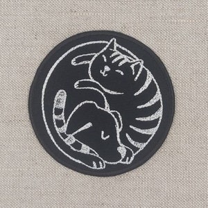 May include: A circular black patch featuring a white embroidered design of a cat and a dog curled together. The patch is set against a neutral, textured background.