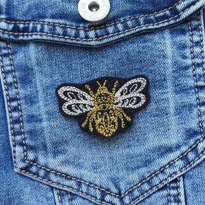 Peut inclure: Gros plan d'une veste en jean avec un patch brodé d'abeille sur la poche. L'abeille est noire, dorée et blanche.